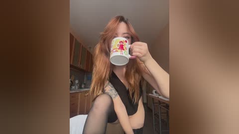 Media: Video of a young woman with long red hair, fair skin, and tattoos on her arms, sitting in a kitchen, holding a ceramic mug with a cartoon character design, wearing a black crop top and black stockings.
