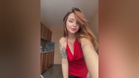 Media: Video of a young Caucasian woman with long, auburn hair and a tattoo on her left arm. She's wearing a red halter top, smiling, and standing in a modern kitchen with light-colored cabinets.