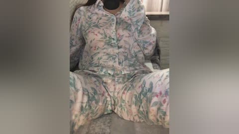 Media: Video of a person seated, wearing floral pajamas with pink, green, and blue hues. Legs spread apart, showing a glimpse of underwear. Background includes a wooden floor and a blurred window.