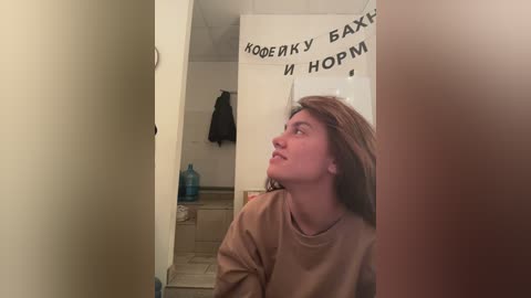 Media: A video captures a young Caucasian woman with long brown hair, wearing a beige sweater, standing in a narrow hallway. A Russian phrase is written on a banner in the background.