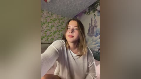 A video of a young Caucasian woman with straight, shoulder-length brown hair, wearing a white long-sleeve shirt, sitting indoors against a mural of a castle. The room has a soft, cozy ambiance.