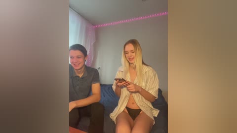 Media: Video of a smiling, light-skinned blonde woman in a white striped shirt, sitting on a dark sofa with a man beside her.