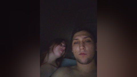 Video of a young man with a beard and a young woman with long hair, both topless, in a dimly lit bedroom.