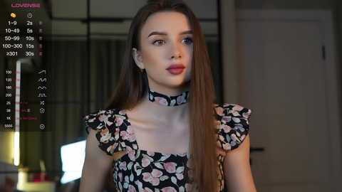 Media: Video of a young woman with long brown hair, wearing a black floral dress and a choker, standing indoors with a modern, dimly lit room in the background.