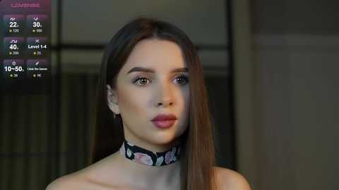 Media: A video of a young woman with fair skin, long brown hair, and a black floral choker, wearing a strapless top. She's in a dimly lit, modern room with a digital interface showing stats in the background.