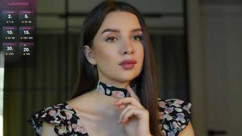 Media: Video of a young woman with fair skin, long dark hair, and light makeup, wearing a floral-patterned top, with a blurred modern indoor background.