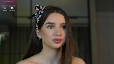 Media: Video of a young Caucasian woman with long brown hair, wearing a floral headband, and light makeup. She has a neutral expression, standing indoors.