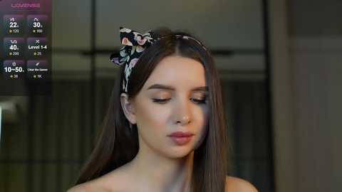 Media: Video of a young woman with fair skin and long brown hair, wearing a black headband with floral print, looking contemplative. Background shows a modern, blurred interior with windows.