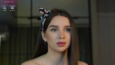 Media: Video of a young woman with long brown hair, wearing a black floral headband, and a soft pink lip color, in a modern interior setting.