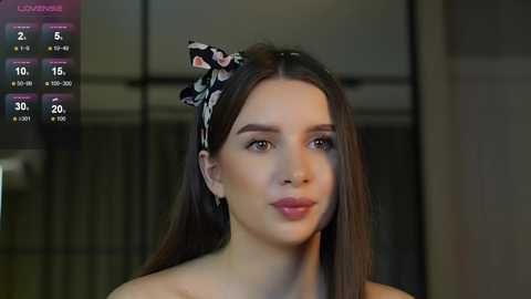 Media: Video of a young woman with fair skin, straight brown hair, and a black floral headband. She has light makeup and wears a strapless top. Background shows a blurred, modern interior with glass doors.