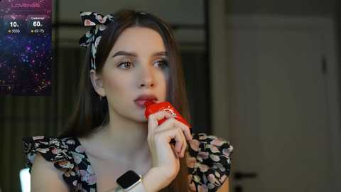 Media: Video of a young woman with fair skin and long brown hair, wearing a floral dress, eating a strawberry while looking thoughtful. Background shows a dimly lit room with a TV displaying a news channel.