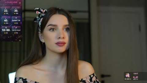 Media: Video of a young woman with long brown hair, wearing a floral off-shoulder top, standing in a dimly lit room.