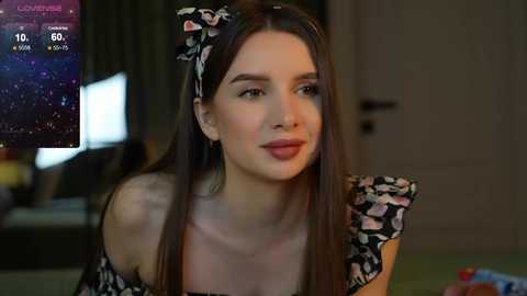 Media: Video of a young woman with long brown hair, wearing a black floral dress and headband, looking serious in a dimly-lit room with a TV showing a nighttime cityscape.