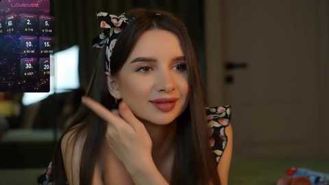 Media: Video of a young woman with long brown hair, wearing a floral headband, in a casual outfit, gesturing with her hand, indoors with a TV displaying a weather forecast.