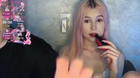 Media: Video of a young woman with light skin and long, pastel pink hair, wearing a black top, applying lipstick. The background shows a textured gray wall with a light switch.