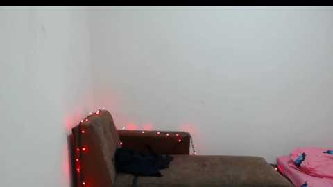 Video of a dimly-lit corner room with a brown sofa, covered in dark fabric, against a plain white wall. Red string lights are wrapped around the sofa's armrests, casting a warm glow. The background is simple and uncluttered, with a hint of a pink blanket visible in the right corner.
