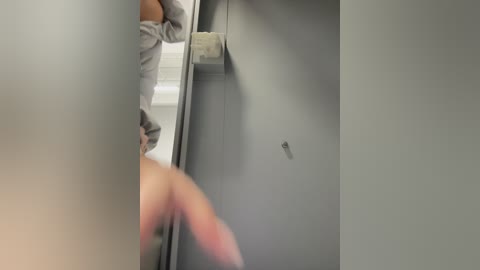Media: Video of a person with light skin, wearing a gray shirt, looking into a mirror, showing a small hole in the wall, and a roll of toilet paper mounted nearby.