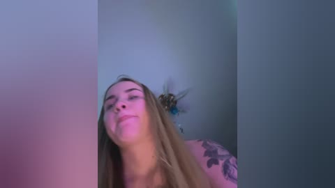 Media: A video of a smiling young woman with long blonde hair, wearing a black top, leaning against a wall with a blue floral tattoo on her arm, bathed in soft, pastel-colored lighting.