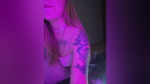 Media: Video of a tattooed woman with long blonde hair, wearing a white tank top, under purple lighting. Her arms display intricate black and gray tattoos. Background is dimly lit, suggesting an indoor setting.