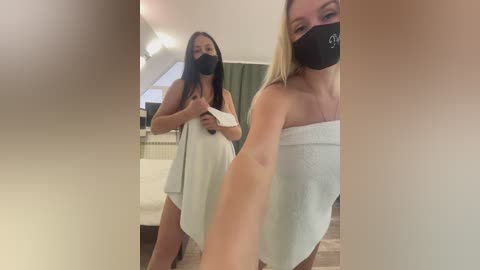 Media: Video of two women wearing black face masks, one holding a towel, in a cozy room with beige carpeting, a green curtain, and a radiator.