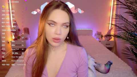 Media: Video of a young, fair-skinned woman with long brown hair and subtle makeup, wearing a pastel pink top, in a cozy bedroom with a plush rug, potted plants, and soft lighting.