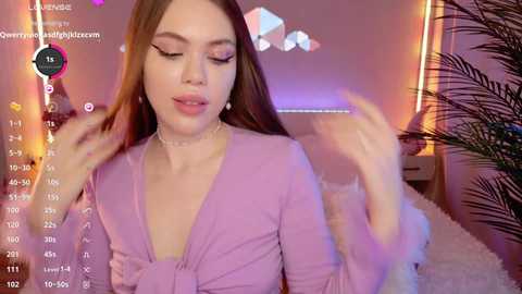 Media: Video of a young woman with long brown hair, wearing a purple dress, smiling and playfully touching her hair, in a cozy, pink-lit bedroom.