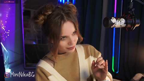 Media: Video of a young woman with light skin, styled hair in two buns, wearing a beige sweater, holding a microphone, set against a dimly lit room with colorful LED lights.