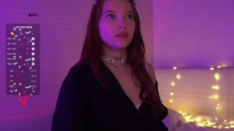Video of a young woman with long, wavy brown hair, wearing a black dress with a white collar, in a softly lit bedroom with purple and yellow lights.