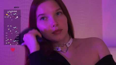 Video of a young Caucasian woman with long brown hair, wearing a black off-shoulder top and a silver choker with a heart pendant. She has light skin and blue eyes, set against a purple-lit background.