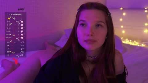 A video of a young woman with long brown hair, wearing a black off-shoulder top, lying on a bed in a dimly lit room with purple lighting and string lights.