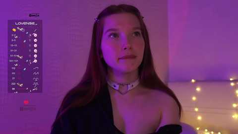 Video of a young woman with long brown hair, wearing a black off-shoulder top, and a white choker. She stands in a dimly lit room with purple lighting, a calendar poster, and fairy lights in the background.