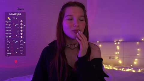 Video of a young woman with long brown hair, wearing a black jacket, sitting on a bed with a purple glow. She's touching her lips with her right hand, and there's a \"LOVEWINS\" poster and string lights in the background.