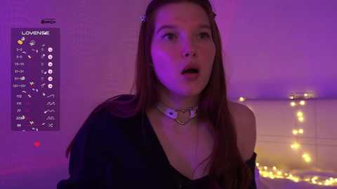 Video of a young woman with long brown hair, wearing a black off-shoulder top and a white choker, against a purple-lit, cozy bedroom background with fairy lights.
