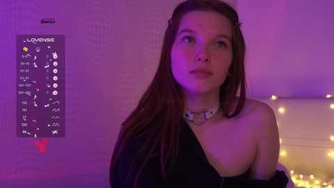 Video of a young woman with long brown hair, wearing a black off-shoulder top, silver choker, and a soft smile. She stands in a dimly lit room with purple lighting, featuring a Lovehoney product chart on the wall.