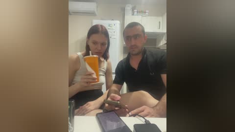 Video of a woman with fair skin and brown hair, dressed in a white tank top, sitting in a kitchen with a man in a black polo shirt. Both are holding smartphones, looking at them. Background includes a refrigerator, microwave, and white cabinets.