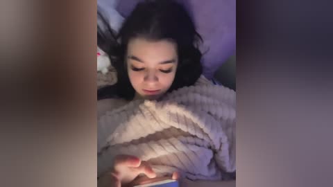 Video of a young girl with dark hair, lying in bed, holding a smartphone, wearing a light-colored, textured blanket. The background is blurred and dimly lit, giving a cozy, intimate atmosphere.