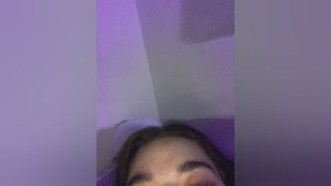 A dimly lit video of a person's face from a low angle, with purple lighting, showing a nose and part of the eyes, against a white wall.