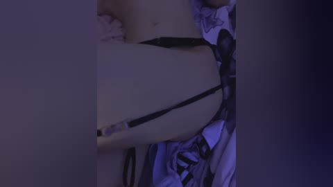 Video of a person wearing a black thong, lying on a bed with a white and gray patterned blanket, dimly lit with a purple hue.