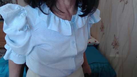 Video of an older woman with fair skin and dark hair wearing a light blue, ruffled blouse and white pants, standing indoors in a room with a floral-patterned curtain and a bed with a blue blanket.