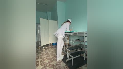 Media: Video of a medical room with a woman in white scrubs and a cap bent over a metal table, surrounded by beige curtains and patterned floor tiles.