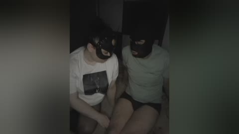 Video of two young women in a dimly lit room, wearing black masks and white T-shirts with a graphic. They sit on the floor, legs crossed, facing each other.