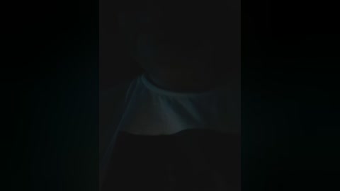 A dimly lit video of a person's head and shoulders, partially obscured by a sheer white fabric draped over the face, creating a veil-like effect. The background is dark, making the fabric stand out.