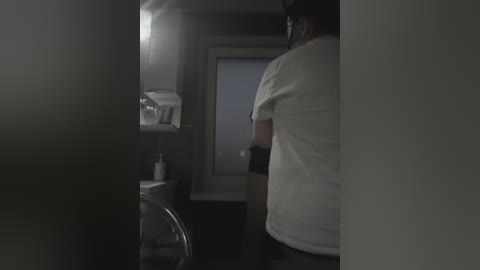 Video of a dimly lit, narrow laundry room with a man, wearing a white T-shirt, standing in the background. The room features a white wall-mounted shelf with a white container and a partially open door with a light source.