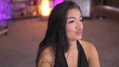Video of a young Asian woman with long black hair, wearing a black halter top, making a pouty face. Background features a blurred bar scene with purple and orange lighting.