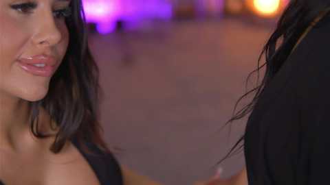 A video captures a close-up of a woman with long, dark hair and a light complexion, smiling. She wears a black top. In the background, blurred purple and orange lights create a vibrant, nightlife atmosphere.