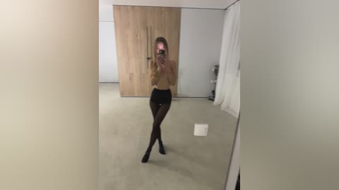A video of a slender, fair-skinned woman with long blonde hair, wearing a beige top, black stockings, and black boots, taking a mirror selfie in a modern, minimalist bedroom with light wood paneling and white curtains.
