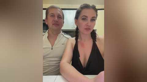 Media: Video of a young couple in a modern kitchen. The man, with short blond hair, wears a white polo shirt. The woman, with long brown hair in braids, dons a black halter top.