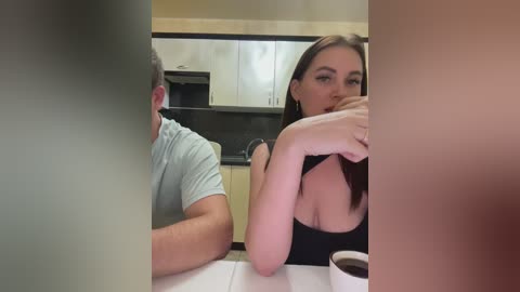 Media: Video of a woman with long brown hair and a large bust, wearing a black top, seated in a kitchen, talking to a man in a light blue shirt, both with blurred faces.