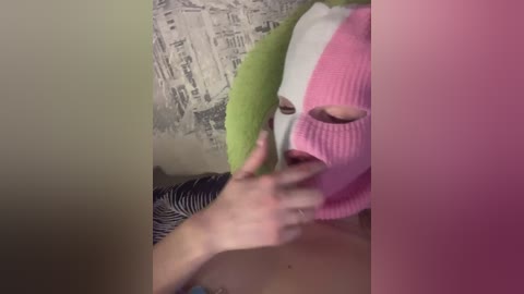 Media: A video of a person with a pink and green balaclava covering their face, hands pulling at it, standing in front of a zebra-print wall.