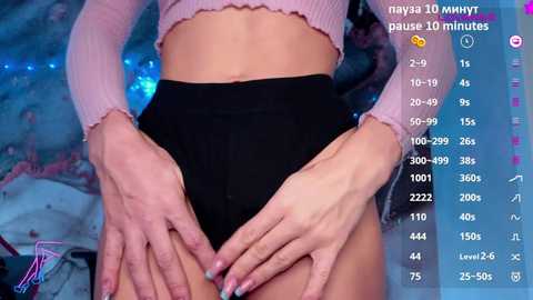 Video of a slender, fair-skinned woman in a pink cropped top and black high-waisted underwear, hands resting on her hips. Background includes a blue-tinted room with soft lighting.